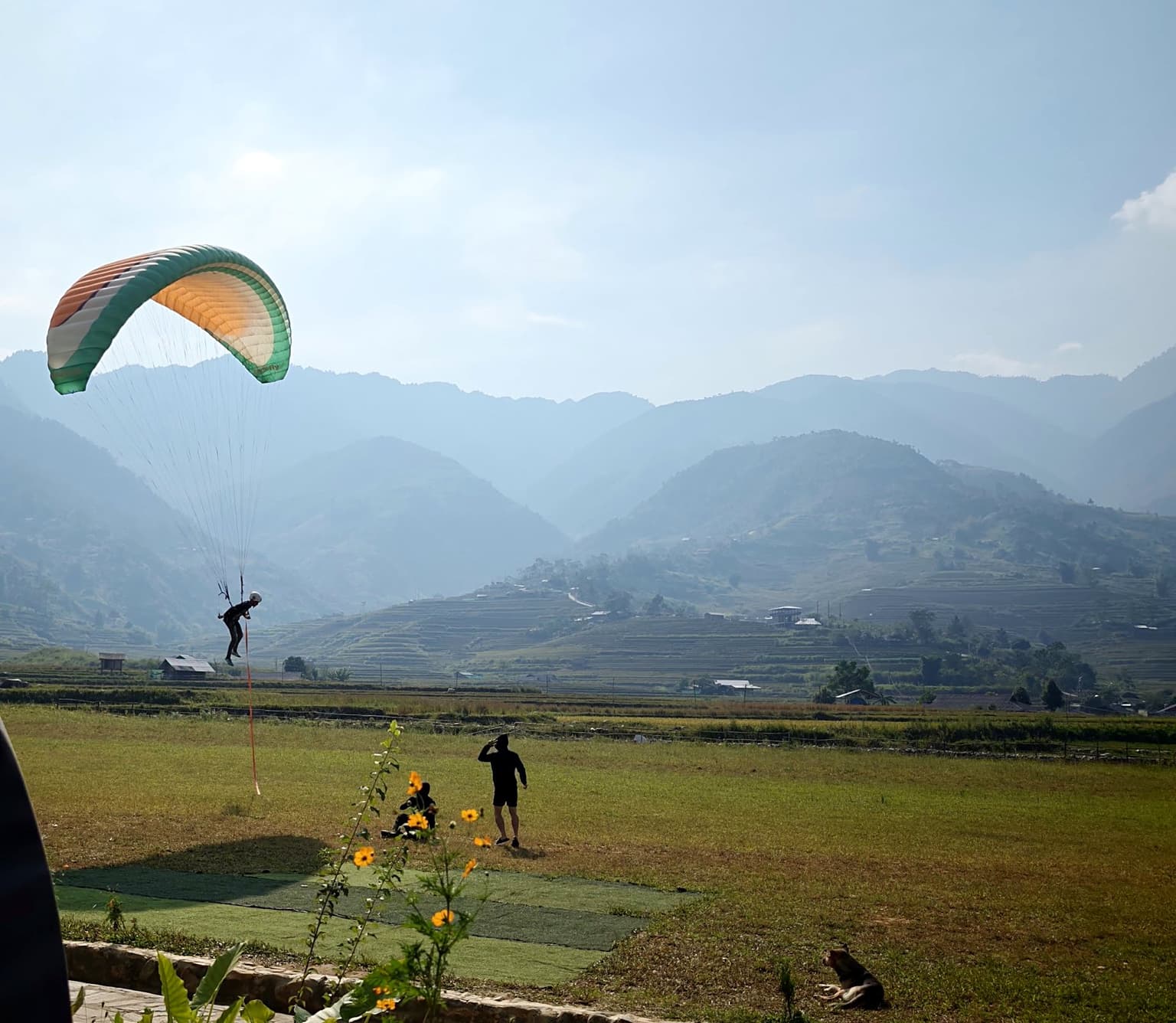 A hand-towed flight: the student is a few metres off the ground, nearly upright, the red tow line running down to the crew; the instructor watches from the field.