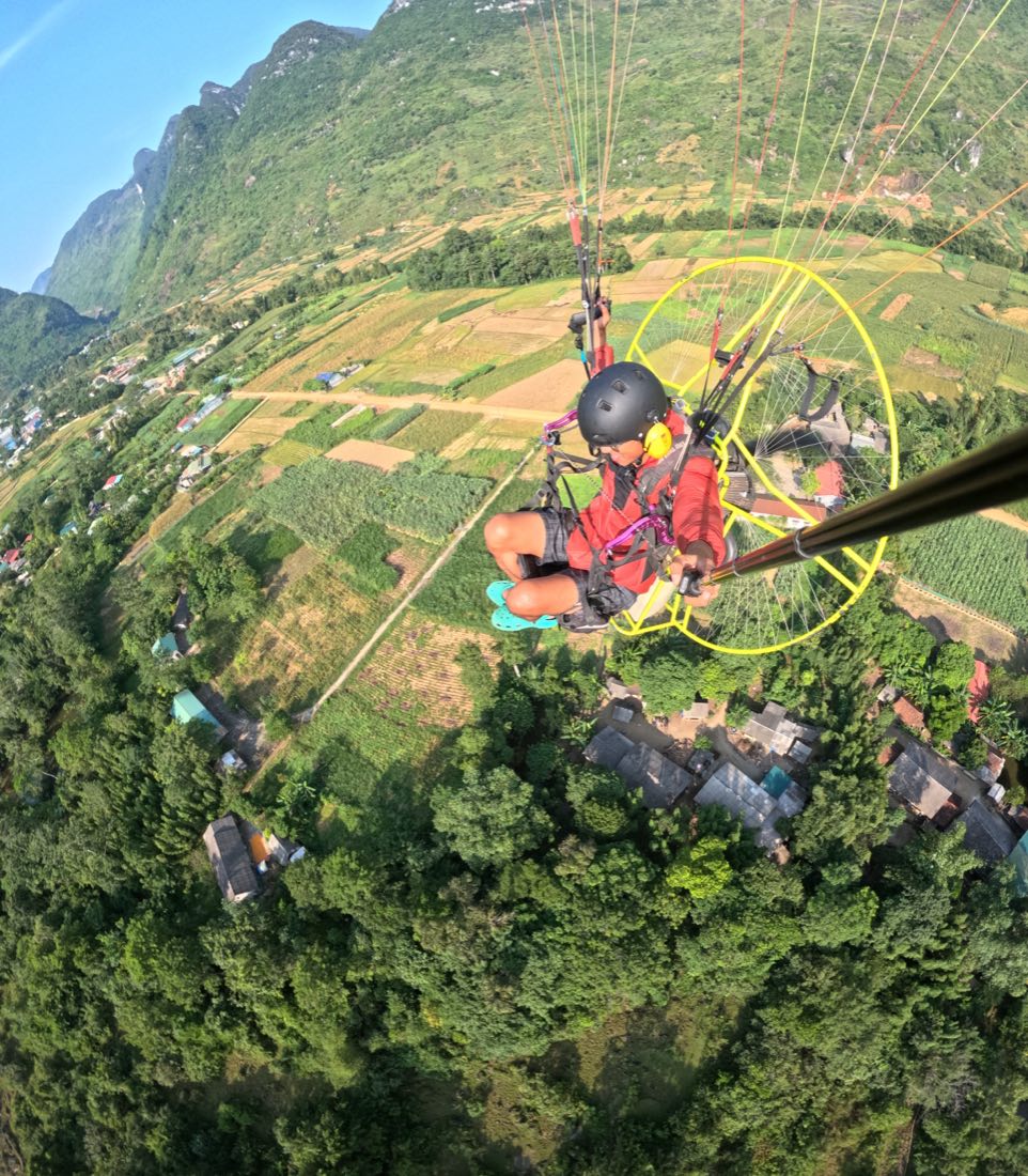 A solo PPG pilot low over fields and a village, with a limestone wall rising right behind. Photo: Mebayluon, Ha Giang, October 2023